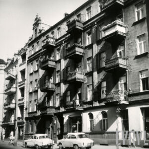 kilkukondygnacyjna kamienica z balkonami, ozdobnymi szczytami i dekoracją architektoniczną na elewacji, przed kamienicą zaparkowane samochody osobowe