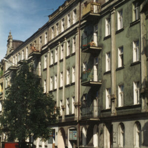 kilkukondygnacyjna kamienica z balkonami, ozdobnymi szczytami i dekoracją architektoniczną na elewacji, przed kamienicą zaparkowane samochody osobowe