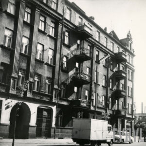 kilkukondygnacyjna kamienica z balkonami, ozdobnymi szczytami i dekoracją architektoniczną na elewacji, przed kamienicą zaparkowane samochody osobowe