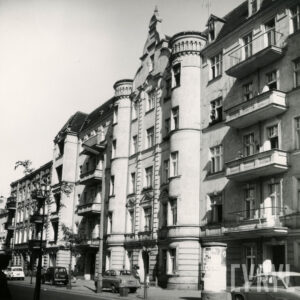 kilkukondygnacyjna kamienica z balkonami, ozdobnymi szczytami i dekoracją architektoniczną na elewacji, przed kamienicą zaparkowane samochody osobowe