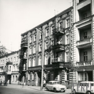 kilkukondygnacyjna kamienica z balkonami, dekoracją architektoniczną na elewacji, przed kamienicą zaparkowane samochody osobowe