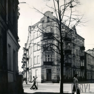 narożna, kilkukondygnacyjna kamienica z balkonami, ozdobnymi szczytami i dekoracją architektoniczną na elewacji, na pierwszym planie drzewo bez liści, chodnikiem spacerują ludzie