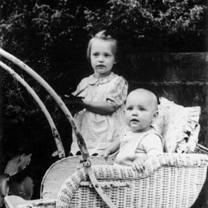 czarno-biała fotografia kilkumiesięcznego dziecka siedzącego w wiklinowym wózku, za nim stoi większa dziewczynka, w tle drzewa
