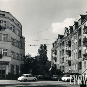 kilkukondygnacyjne budynki po obu stronach ulicy, z prawej wzdłuż chodnika zaparkowane samochody