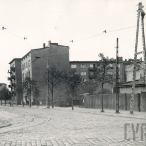 brukowana ulica z torami tramwajowymi, w głębi kilkukondygnacyjne budynki mieszkalne, przed nimi, z prawej drewniane ogrodzenie