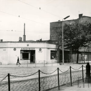 niski narożny budynek z wejściem na narożniku, na pierwszym planie brukowana ulica z torami tramwajowymi, chodnik odgrodzony od ulicy łańcuchami