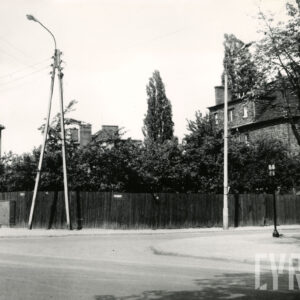 skrzyżowanie ulic, na narożniku pusta działka ogrodzona drewnianym płotem, z prawej fragment nieotynkowanego domu mieszkalnego