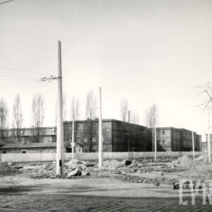 na pierwszym planie brukowana ulica, za nią ulica w czasie budowy, w głębi budynki zakładu, słupy z trakcją tramwajową