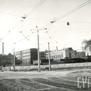 na pierwszym planie brukowana ulica z torami tramwajowymi, w głębi budynki zakładu, słupy z trakcją tramwajową