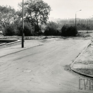 gruntowa ulica, z lewej niski murek, obniżający się wraz z terenem, przy chodnikach betonowe słupy latarni ulicznych, w głębi krzaki i niskie drzewa