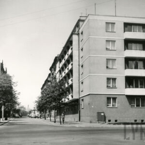 widok na ulicę obsadzoną drzewami, przy niej budynki mieszkalne, z prawej, na narożniku pięciopiętrowy blok mieszkalny z balkonami, w części, krótsza elewacja czteropiętrowa z balkonami