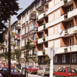 kilkukondygnacyjne budynki mieszkalne wzdłuż ulicy, przy krawężniku zaparkowane samochody