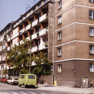 pięciopiętrowy blok mieszkalny z balkonami na narożniku ulic, przy krawężniku zaparkowane samochody osobowe, na pierwszym planie przejście dla pieszych
