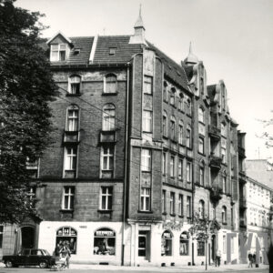 czteropiętrowa kamienica z zamieszkałym poddaszem i hełmami na dachu, zaparkowany samochód, przez ulicę przechodzi kobieta z dziecięcym wózkiem