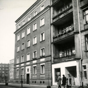 czteropiętrowy budynek mieszkalny, z prawej fragment kilkupiętrowej kamienicy z balkonami, chodnikiem idą ludzie