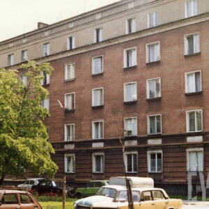 czteropiętrowy budynek mieszkalny, przed nim fragment skweru, wokół niego zaparkowane samochody