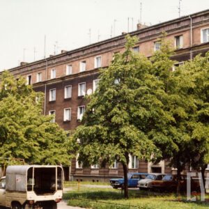 czteropiętrowy budynek mieszkalny przysłonięty przed drzewa rosnące na skwerze, zaparkowane samochody