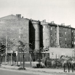 kilkukondygnacyjna kamienica, nieotynkowana, przed nią drewniane ogrodzenie i niezabudowana działka z niskimi drzewami