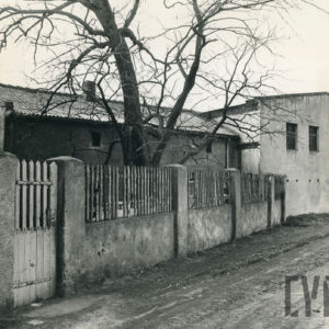 parterowy dom ze spadzistym dachem otoczony murowano-drewnianym płotem ustawionym wzdłuż szutrowej ulicy