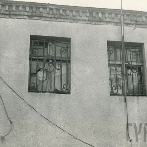 dwa zakratowane okna otynkowanego domu mieszkalnego, nad nimi ozdobny, ceglany gzyms