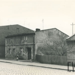 trzy domy ustawione kalenicami do wybrukowanej drogi: piętrowy ceglany, parterowy z poddaszem i parterowy ze spadzistym dachem otoczony drewnianym płotem