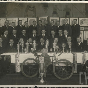 grupa mężczyzn w garniturach siedząca i stojąca na tle dyplomów, przed stołem, na którym ustawione są puchary i nagrody; na pierwszym planie przed nimi stoi rower, a obok niego mały chłopiec