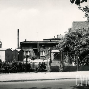 w głębi parterowy, murowany budynek, z lewej trzy garaże, całość ogrodzona metalową siatką z bramą i furtką, na posesji drzewa i krzewy, w głębi z lewej wysoki murowany komin