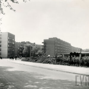 asfaltowa ulica z chodnikiem, w głębi po lewej i prawej stronie ulicy kilkukondygnacyjne bloki mieszkalne, chodnikiem i ulicą spacerują ludzie