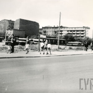 na pierwszym planie asfaltowa ulica, chodnikiem spacerują ludzie, dalej nieogrodzony teren z ułożonymi na nim rurami, w głębi wysokie kamienice, widoczne ściany pozbawione okien, z prawej  kilkukondygnacyjny blok mieszkalny