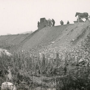 na pierwszym planie nieużytki, w głębi skarpa, na niej koń z wozem i grupa ludzi przy pracach ziemnych