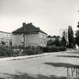 brukowana ulica, przy niej kilkukondygnacyjne domy mieszkalne w ogrodach, ogrodzone, w głębi wysoki blok mieszkalny