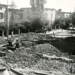 brukowana ulica, przy niej kilkukondygnacyjne domy mieszkalne w ogrodach, ogrodzone, na pierwszym planie rozkopana ulica, montaż rur