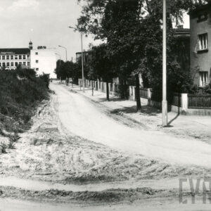 brukowana ulica, przy niej kilkukondygnacyjne domy mieszkalne w ogrodach, ogrodzone, z lewej skarpa, za nią kilkukondygnacyjny budynek, za nim wysoka wieża