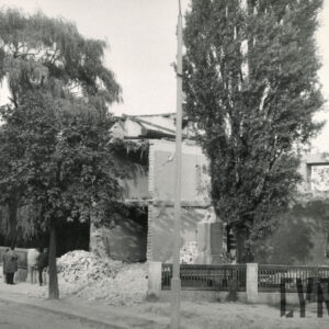 kilkukondygnacyjny budynek mieszkalny w trakcie rozbiórki, wokół budynku gruz, drzewa, całość otoczona drewniano-murowanym ogrodzeniem