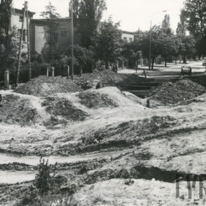 na pierwszym planie rozkopana ulica, przy niej za drzewami kilkukondygnacyjne domy mieszkalne w ogrodach, ogrodzone