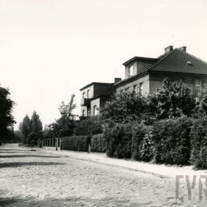 brukowana ulica, przy niej kilkukondygnacyjne domy mieszkalne w ogrodach, ogrodzone