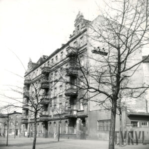 czteropiętrowa kamienica z ozdobnymi szczytami, na pierwszym planie ulica, przy chodnikach drzewa bez liści