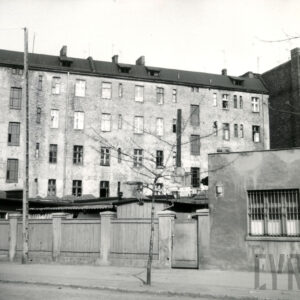 na pierwszym planie za ogrodzeniem niskie budynki, w głębi kilkukondygnacyjna, prosta elewacja kamienicy