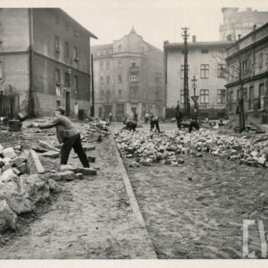 prace brukarskie na ulicy, na środku ulicy i po bokach leżą zgrupowane kostki brukowe, z lewej jeden mężczyzna rzuca do drugiego kostkę brukową, w głębi pochyleni mężczyźni, po obu stronach ulicy i na wprost kilkukondygnacyjne budynki i kamienice