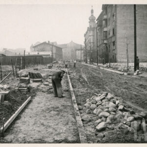 prace brukarskie na ulicy, na środku ulicy i po bokach leżą zgrupowane kostki brukowe, na środku pochylony mężczyzna, po prawej stronie ulicy i na wprost kilkukondygnacyjne budynki i kamienice, z lewej drewniane ogrodzenie