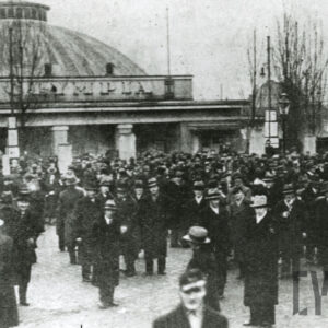 duża grupa ludzi, w większości mężczyźni w płaszczach, garniturach i kapeluszach na głowie stojący na tle budynku z dużą kopułą