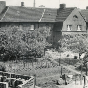 dwukondygnacyjny budynek mieszkalny z ryzalitem i spadzistym dachem, przed nim ogródki z niskimi drzewkami