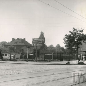 na pierwszym planie brukowana ulica z torami tramwajowymi, w głębi niskie budynki gospodarcze ogrodzone metalową siatką, za nimi dwukondygnacyjny budynek mieszkalny ze spadzistym dachem, z prawej przystanek tramwajowy, na nim czekają ludzie na tramwaj