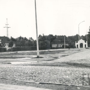 na pierwszym planie brukowana ulica, w głębi zadrzewiony teren, przed nim jednokondygnacyjny budynek ze spadzistym dachem i strychem, z jego prawej strony ozdobna brama wjazdowa