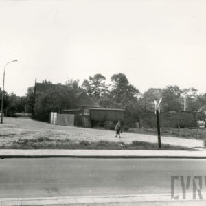 na pierwszym planie asfaltowa ulica z chodnikami, w głębi ogrodzony teren z budynkami gospodarczymi