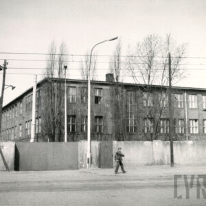 trzykondygnacyjny, ceglany budynek fabryczny z dużymi oknami, ogrodzony murowany płotem z metalową bramą, na pierwszym planie brukowana ulica, chodnikiem idzie młody mężczyzna