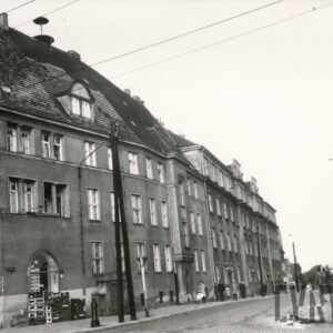 brukowana ulica, po jej lewej stronie kilkukondygnacyjne domy mieszkalne ze spadzistym dachem, strychem