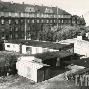 na pierwszym planie drewniane szopki, garaże i niskie budynki gospodarcze, w głębi czterokondygnacyjny budynek ze spadzistym dachem, z prawej niższy budynek mieszkalny z trójkątnymi szczytami