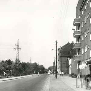 asfaltowa ulica, po obu jej stronach chodniki, z prawej kiosk handlowy, przy nim nachylona kobieta, dalej kilkukondygnacyjne budynki mieszkalne