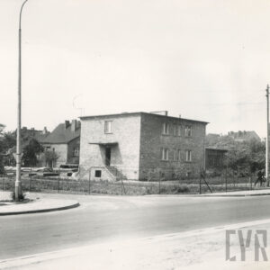 skrzyżowanie ulic, za nim dwukondygnacyjny budynek mieszkalny z płaskim dachem z ogródkiem ogrodzonym metalową siatką